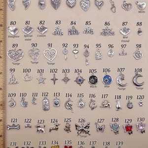 May include: A collection of silver-toned metal charms numbered 70 through 139, featuring hearts, stars, moons, animals, and insects. A wooden ruler on the left shows measurements in inches. Some charms include text like Mom, Sister, and Love.