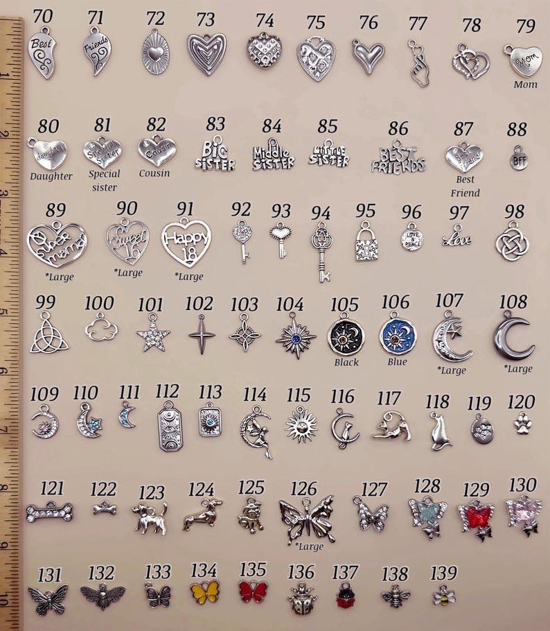 May include: A collection of silver-toned metal charms numbered 70 through 139, featuring hearts, stars, moons, animals, and insects. A wooden ruler on the left shows measurements in inches. Some charms include text like Mom, Sister, and Love.