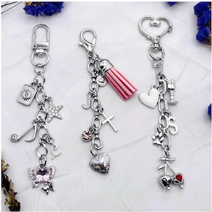 May include: Three silver-toned metal keychains featuring various charms. The first has a tarot card, star, music note, letter L, and pink butterfly. The second has a pink tassel, letter J, cross, and heart locket. The third has a heart, goblet, letter S, paw print, and cherries.