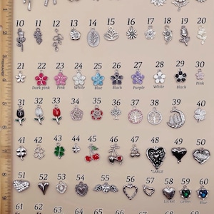 May include: A collection of 69 silver-toned metal charms arranged in rows, numbered 1 through 69. The charms feature various shapes including bows, flowers, trees, hearts, and fruits, with some colored enamel accents. A 10-inch ruler is visible on the left.