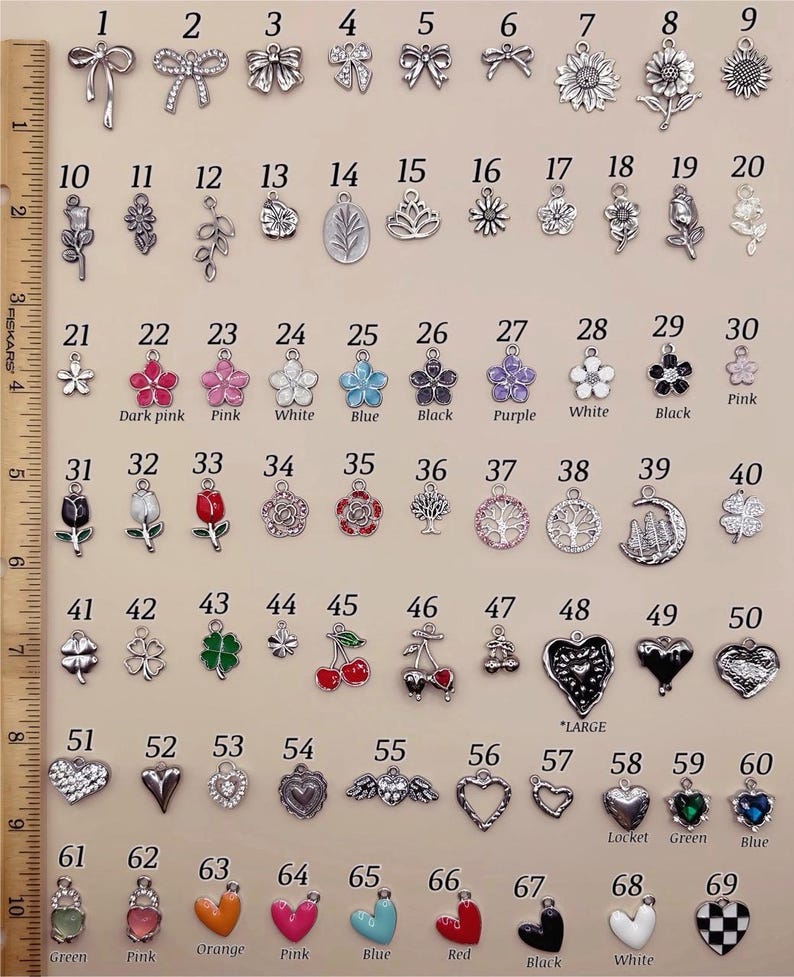 May include: A collection of 69 silver-toned metal charms arranged in rows, numbered 1 through 69. The charms feature various shapes including bows, flowers, trees, hearts, and fruits, with some colored enamel accents. A 10-inch ruler is visible on the left.
