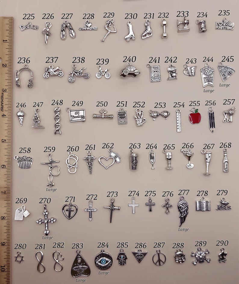 May include: A collection of silver-toned metal charms arranged in rows, numbered 225 through 290, featuring various shapes including sports, food, and symbols. A wooden ruler on the left shows measurements in inches for scale reference.