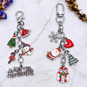 May include: Two silver-toned metal keychains featuring assorted Christmas-themed charms. The left keychain includes a snowman, a candy cane, a Christmas tree, a Santa hat, and a Merry Christmas sign. The right keychain features a snowflake, a heart, a Santa Claus, a Christmas tree, and a gift box.