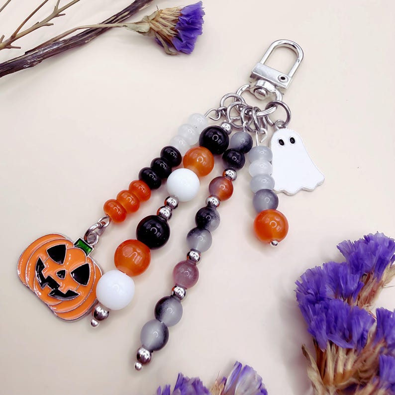 Puede incluir: Un llavero con un cierre de mosquet&oacute;n plateado y m&uacute;ltiples hilos de cuentas colgantes en naranja, negro, blanco y gris. Los hilos est&aacute;n decorados con un dije de calabaza de metal y un dije de fantasma blanco, creando un accesorio festivo.