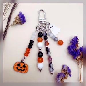 May include: A Halloween-themed keychain featuring a silver-toned lobster clasp and three dangling beaded strands. The strands include orange, black, white, and grey beads, finished with a metal jack-o'-lantern charm and a white ghost charm.