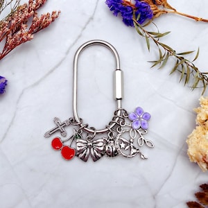 May include: A silver-toned oval carabiner keychain featuring multiple dangling charms, including a silver cross, two red cherries with a green leaf, a silver bow, a ladybug, a crescent moon with a star, and a purple five-petaled flower.