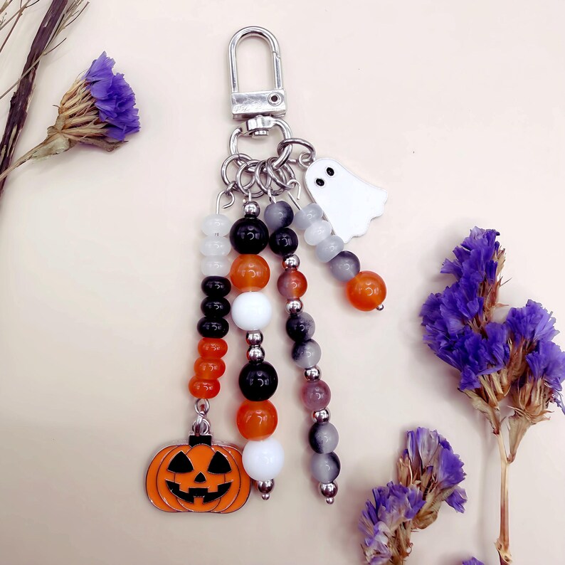 Puede incluir: Un llavero con un cierre de mosquet&oacute;n plateado y tres hilos colgantes de cuentas en naranja, negro, blanco y gris. Los hilos est&aacute;n adornados con un dije de calabaza de metal y un peque&ntilde;o dije de fantasma blanco.