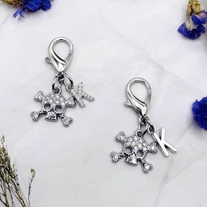 May include: Two silver-toned charms with lobster clasps. Each charm features a rhinestone-studded skull and a rhinestone-covered letter "K". The charms are designed for jewelry making or as decorative accessories.
