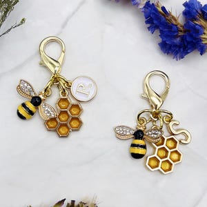 May include: Two gold-colored bee-themed charms with lobster clasps. Each charm features a bee with yellow and black stripes, rhinestone wings, and a honeycomb. One charm has a white circle with the letter "R", the other has the letter "S".