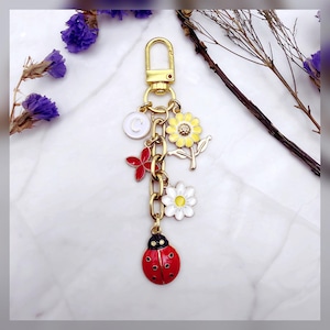 May include: A gold-toned keychain with a lobster clasp, featuring a chain and several charms. Charms include a white circle with a "C", a red flower, a yellow sunflower, a white daisy, and a red ladybug with black spots. The background is a marble-like surface.