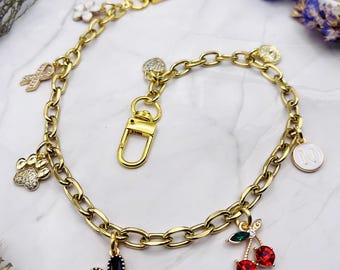 May include: A gold-toned chain bracelet featuring multiple charms including a black heart, red crystal cherries, a paw print with clear stones, a ribbon, a white flower, a heart, and a circular pendant with the letter N in white enamel.