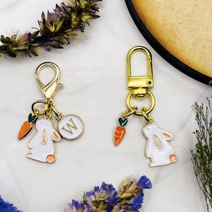 May include: Two gold keychains with charms. One keychain has a white rabbit charm with a carrot and a green heart. The other keychain has a white rabbit charm with a carrot and a green heart, and a round charm with the letter W.