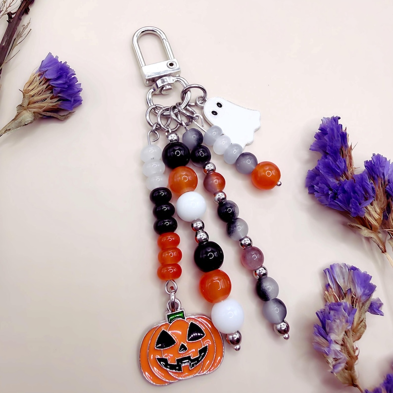 Puede incluir: Un llavero con un cierre de mosquet&oacute;n plateado, un dije de fantasma blanco y un dije de calabaza de esmalte naranja con detalles negros. El dise&ntilde;o incluye tres hilos colgantes de cuentas en colores blanco, negro, naranja y gris transl&uacute;cido.