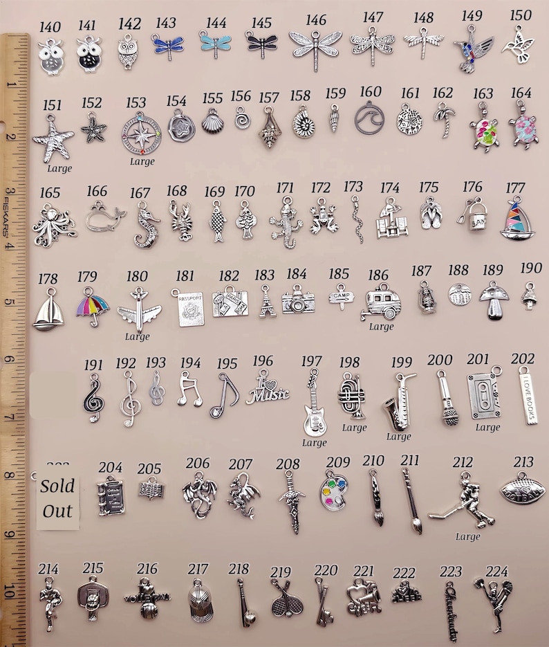 May include: A collection of silver-toned metal charms arranged in rows, numbered 140 through 224, featuring various shapes like animals, musical instruments, and sports gear, displayed next to a wooden ruler measuring 10 inches in length.