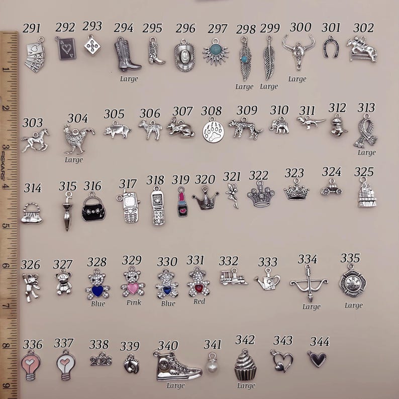 May include: A collection of silver-toned metal charms arranged in rows, numbered 291 through 344, featuring various shapes including animals, objects, and symbols. A wooden ruler on the left shows measurements in inches for scale reference.