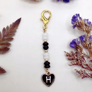 May include: A beaded charm featuring a gold-tone lobster clasp, alternating white and black disc-shaped beads, and a heart-shaped pendant with a black enamel background and a silver-tone letter H centered on the front of the heart charm.