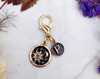 Puede incluir: Un dije con cierre de mosquetón de metal dorado que incluye dos colgantes circulares. Uno muestra un fondo de esmalte negro con un motivo de sol y luna dorado y un pequeño cristal ámbar. El segundo colgante más pequeño presenta la letra P en negro.