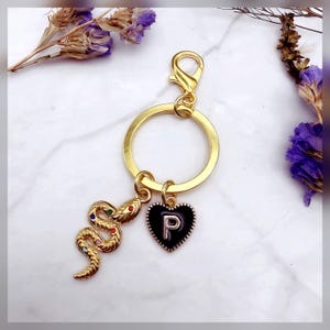 May include: Gold-toned keychain with a large ring and lobster clasp. The keychain features a gold snake charm with colorful gem accents and a black heart charm with the letter "P". The background includes dried flowers.