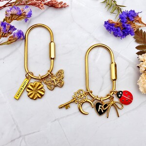 May include: Two gold-toned oval carabiner keychains. The left keychain features a rectangular charm with the text I LOVE BOOKS, a flower, and a butterfly. The right keychain features a key, a crescent moon, a black heart with the letter R, a bow, and a ladybug.