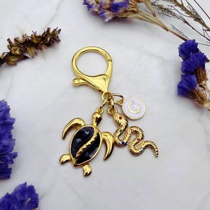 May include: Gold-colored keychain with a lobster clasp, featuring a turtle charm with a black enamel shell and a snake charm adorned with colorful gemstones. A white circular charm with a letter "C" is also included.