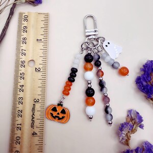 Puede incluir: Un llavero con tem&aacute;tica de Halloween que incluye un cierre de mosquet&oacute;n plateado y tres hilos de cuentas. Los hilos tienen cuentas naranjas, negras, blancas y grises, con un dije de calabaza y un fantasma blanco. Una regla muestra 10 cm.
