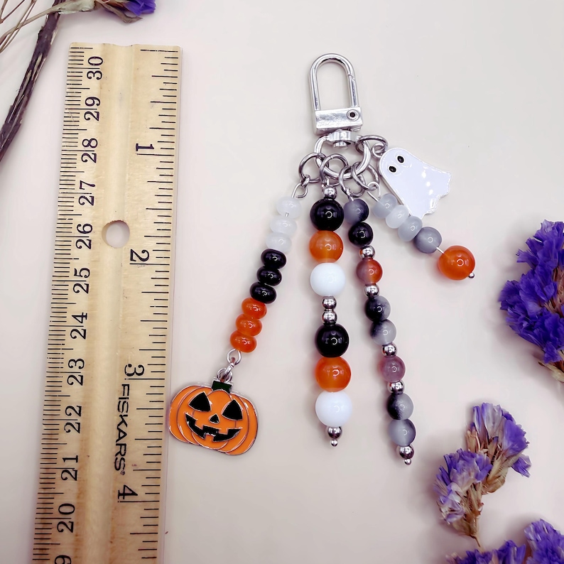 Puede incluir: Un llavero con tem&aacute;tica de Halloween que incluye un cierre de mosquet&oacute;n plateado y tres hilos de cuentas. Los hilos tienen cuentas naranjas, negras, blancas y grises, con un dije de calabaza y un fantasma blanco. Una regla muestra 10 cm.