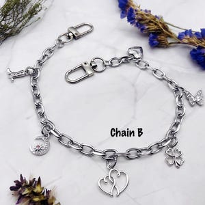 May include: A silver-toned metal chain bracelet labeled Chain B, featuring multiple charms including a cowboy boot, a crescent moon with a pink stone, two interlocking hearts, a four-leaf clover, and a butterfly, secured with two lobster clasps.