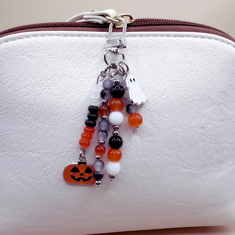 Puede incluir: Un llavero de metal plateado con tres hileras de cuentas colgantes en naranja, negro, blanco y gris transl&uacute;cido. El llavero incluye un dije de calabaza naranja plano y un dije de fantasma blanco, sujeto a un bolso de color blanco y marr&oacute;n.