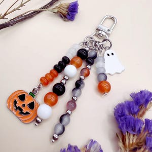 Puede incluir: Un llavero con un cierre de mosquet&oacute;n plateado y m&uacute;ltiples hilos de cuentas colgantes en naranja, negro, blanco y gris. Los hilos est&aacute;n decorados con un dije de calabaza de metal y un dije de fantasma blanco, creando un accesorio festivo.