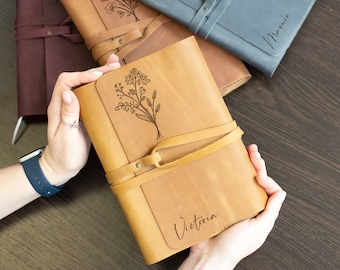 May include: Three leather-bound journals with floral designs and personalized names. The journals are brown, blue, and tan. The tan journal has the name "Victoria" engraved on the front.