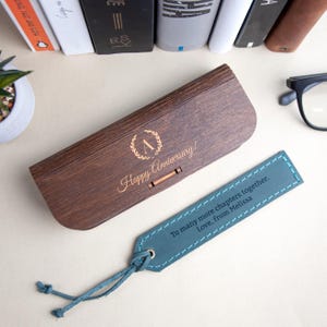 Może przedstawiać: Drewniane etui na okulary z spersonalizowanym napisem "Happy Anniversary!" i niebieską sk&oacute;rzaną zakładką z tekstem "To many more chapters together. Love, from Melissa."