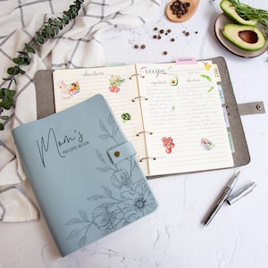 May include: A light blue recipe book with a floral design and the text "Mom's Recipe Book" on the cover. The book is open to a page with lined paper and a section for ingredients, directions, and notes.