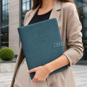 Op de afbeelding: Een blauwgroene documentenmap met een gladde, matte afwerking en een zijsluiting met drie koperkleurige metalen studs. De voorkant is voorzien van een in reliëf aangebrachte florale lijntekening en de tekst Naomi Brown Real Estate Agent.