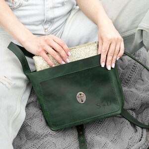 K&ouml;nnte beinhalten: Eine dunkelgr&uuml;ne Umh&auml;ngetasche aus Leder mit einer Vordertasche, einem metallischen Drehverschluss und den eingravierten Buchstaben S und D mit einem kleinen Blumen-Symbol. Eine goldene Glitzer-Tasche steckt teilweise im Hauptfach.