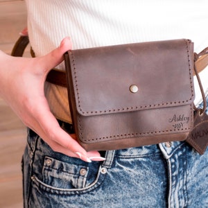May include: A brown leather fanny pack with a flap closure and a silver snap button. The fanny pack has the name "Ashley" engraved on the front.