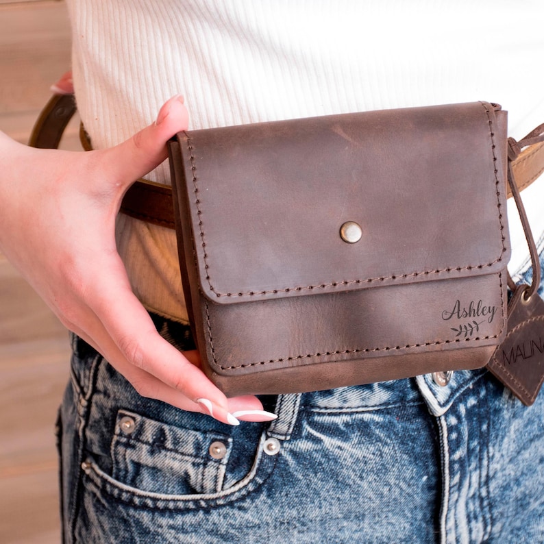 May include: A brown leather fanny pack with a flap closure and a silver snap button. The fanny pack has the name "Ashley" engraved on the front.