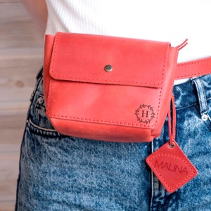 May include: A red leather fanny pack with a small, square leather tag that says "MALINA". The fanny pack has a metal snap closure and a small, engraved circle with the letter "H" inside a wreath.