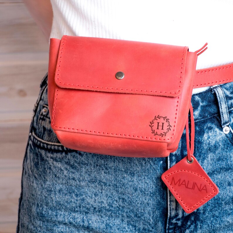 May include: A red leather fanny pack with a small, square leather tag that says "MALINA". The fanny pack has a metal snap closure and a small, engraved circle with the letter "H" inside a wreath.