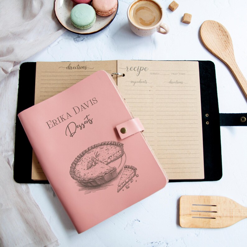 a pink recipe book with a pie illustration, a wooden spoon, a cup of coffee, and a plate of macarons on a white marble surface.