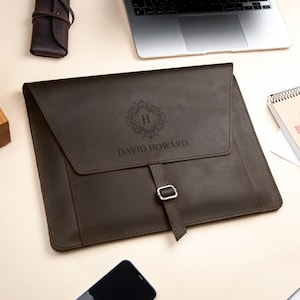 Può includere: Una custodia per laptop in pelle marrone con un design monogrammato. La custodia ha una patta con chiusura a fibbia e le iniziali "H" in una ghirlanda decorativa con il nome "DAVID HOWARD" inciso sotto.