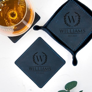 May include: Four blue leather coasters with stitched edges. Each coaster features a monogrammed initial "W" inside a wreath with the name "WILLIAMS" and the year "EST. 2023" engraved in black.