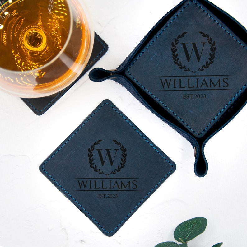 May include: Four blue leather coasters with stitched edges. Each coaster features a monogrammed initial "W" inside a wreath with the name "WILLIAMS" and the year "EST. 2023" engraved in black.