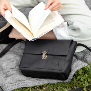 K&ouml;nnte beinhalten: Eine schwarze Umh&auml;ngetasche aus Leder mit rechteckiger Klappe, einem bronzefarbenen Metall-Druckverschluss und den eingepr&auml;gten Buchstaben VA auf der Vorderseite. Die Tasche verf&uuml;gt &uuml;ber sichtbare N&auml;hte an den Klappenr&auml;ndern und einen d&uuml;nnen Schulterriemen.