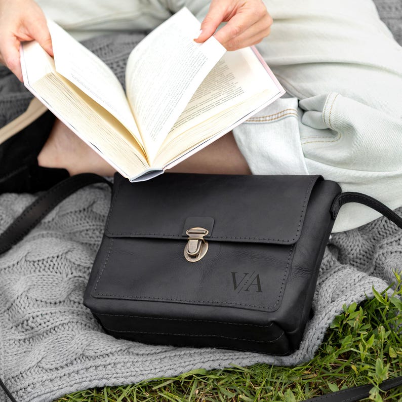 K&ouml;nnte beinhalten: Eine schwarze Umh&auml;ngetasche aus Leder mit rechteckiger Klappe, einem bronzefarbenen Metall-Druckverschluss und den eingepr&auml;gten Buchstaben VA auf der Vorderseite. Die Tasche verf&uuml;gt &uuml;ber sichtbare N&auml;hte an den Klappenr&auml;ndern und einen d&uuml;nnen Schulterriemen.