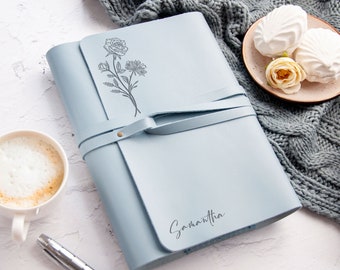 May include: A light blue leather journal with a floral design and the name Samantha embossed on the cover. The journal has a strap closure and is lying on a white surface.