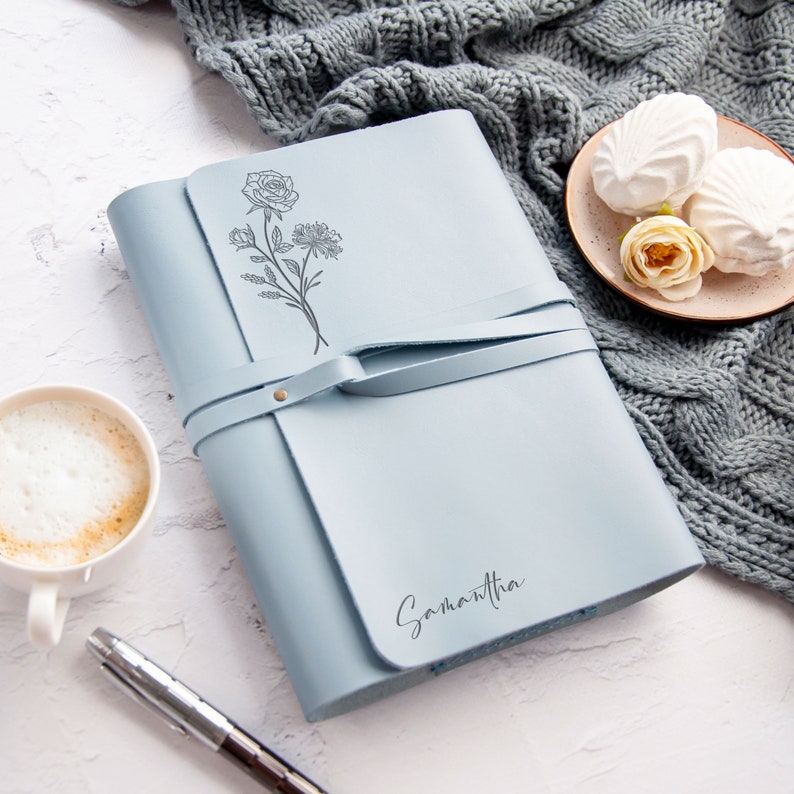 May include: A light blue leather journal with a floral design and the name Samantha embossed on the cover. The journal has a strap closure and is lying on a white surface.