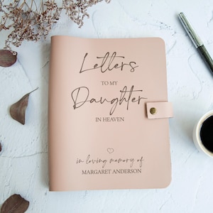 May include: A pale pink leather journal with the words "Letters to my Daughter in Heaven" inscribed in a stylish font. The journal also features the phrase "in loving memory of Margaret Anderson" and a heart symbol. A pen and a coffee cup are visible.