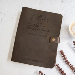 K&ouml;nnte beinhalten: Ein dunkelbraunes Lederjournal mit der Aufschrift "Letters to my little boy as I watch you grow" und dem Namen "Oliver Anderson". Das Journal hat einen Lederriemen mit einem goldenen Knopfverschluss.