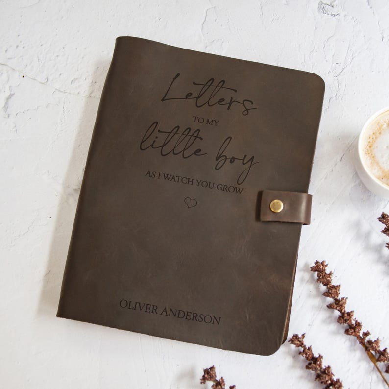 K&ouml;nnte beinhalten: Ein dunkelbraunes Lederjournal mit der Aufschrift "Letters to my little boy as I watch you grow" und dem Namen "Oliver Anderson". Das Journal hat einen Lederriemen mit einem goldenen Knopfverschluss.