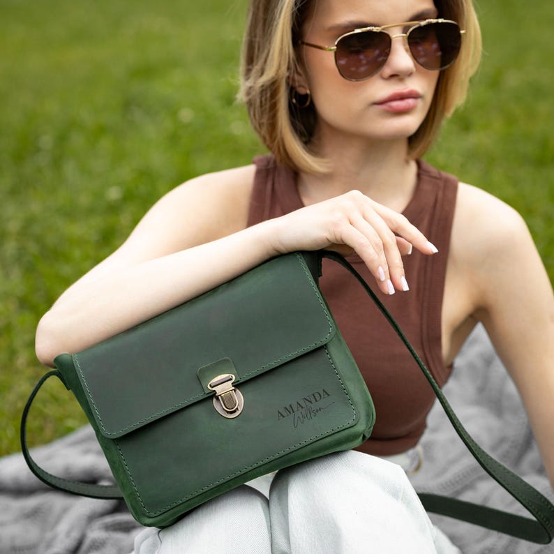 K&ouml;nnte beinhalten: Eine dunkelgr&uuml;ne Umh&auml;ngetasche aus Leder mit &Uuml;berschlag, einem metallischen Druckknopfverschluss und einem d&uuml;nnen Schulterriemen. Auf der Vorderseite der Tasche ist der Name AMANDA WILSON in einer dunklen, eleganten Schreibschrift eingraviert.